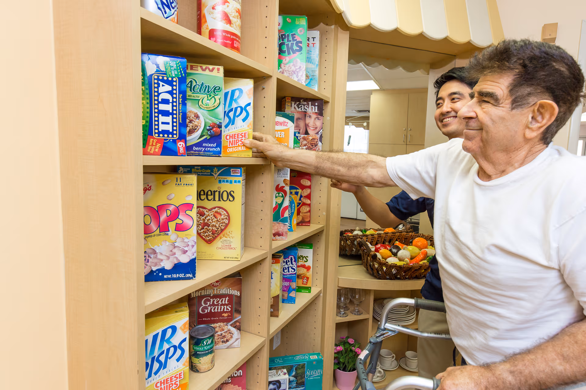 An elderly man using a walker is reaching for a box of cereal on a shelf in a pantry or kitchen area, assisted by a younger man standing behind him. The shelves are stocked with various cereal boxes and other food items, and there is a basket of fruit on the counter in the background.