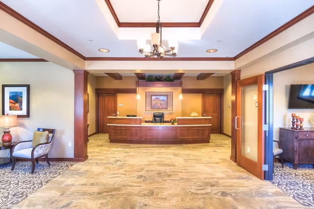 Reception area of Catalina Springs Memory Care featuring a wooden front desk with a chair behind it, two pendant lights hanging above, and a framed picture on the wall. The space has beige walls, tiled flooring, and a chandelier on the ceiling. To the left, there is a small seating area with a chair, side table, and lamp. To the right, there is a room with a glass door, a TV mounted on the wall, and a wooden cabinet with decorative items.