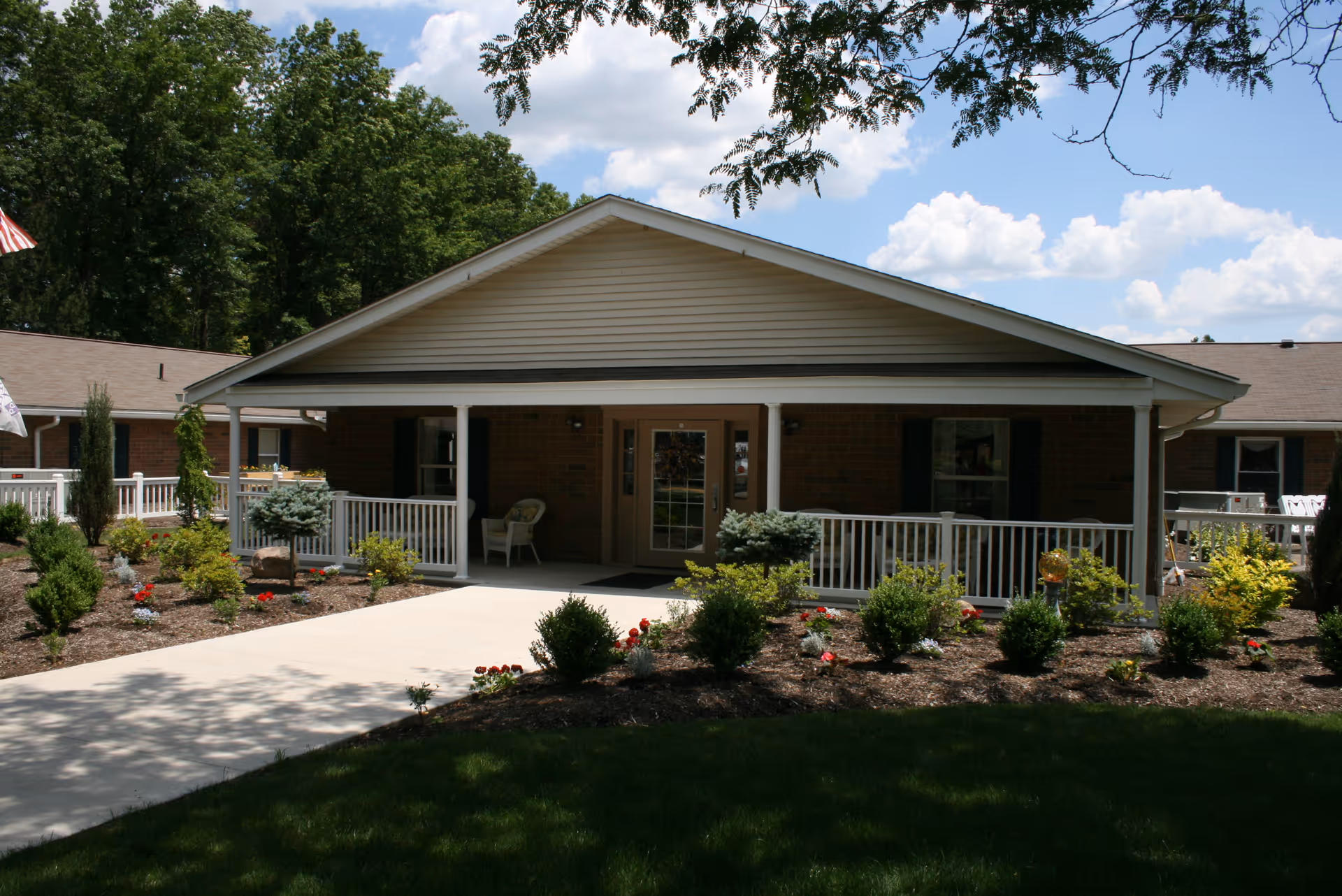 Front exterior view of a single-story brick building with a covered porch, white railing, and a concrete walkway leading to the entrance. The surrounding area features landscaped bushes, small trees, and flowers under a partly cloudy sky.
