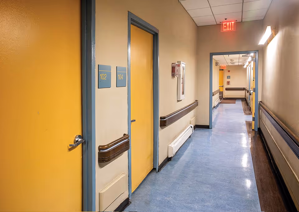 A clean, well-lit hallway in a senior living facility with yellow doors labeled 102 and 104, blue flooring, beige walls, handrails on both sides, and an illuminated exit sign at the end of the corridor.