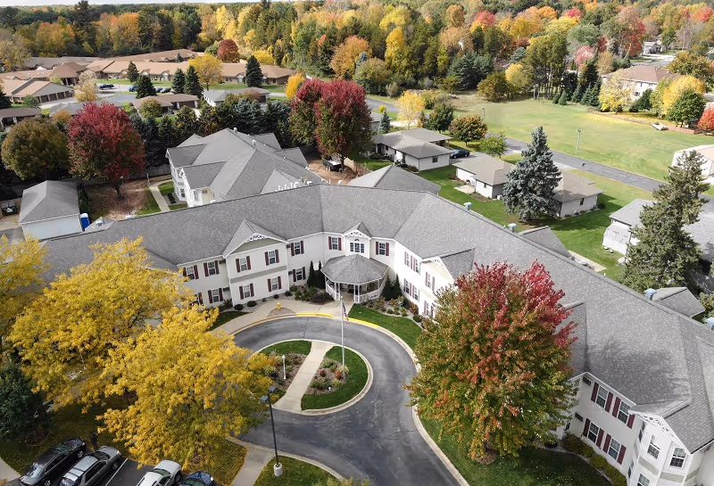 Aerial view of a senior living facility named Renaissance by Rennes - Marinette surrounded by trees with autumn foliage. The building is large with a gray roof and white exterior walls with red shutters. There is a circular driveway in front of the main entrance, and several cars are parked nearby. The surrounding area includes other buildings and a mix of green and colorful fall trees.