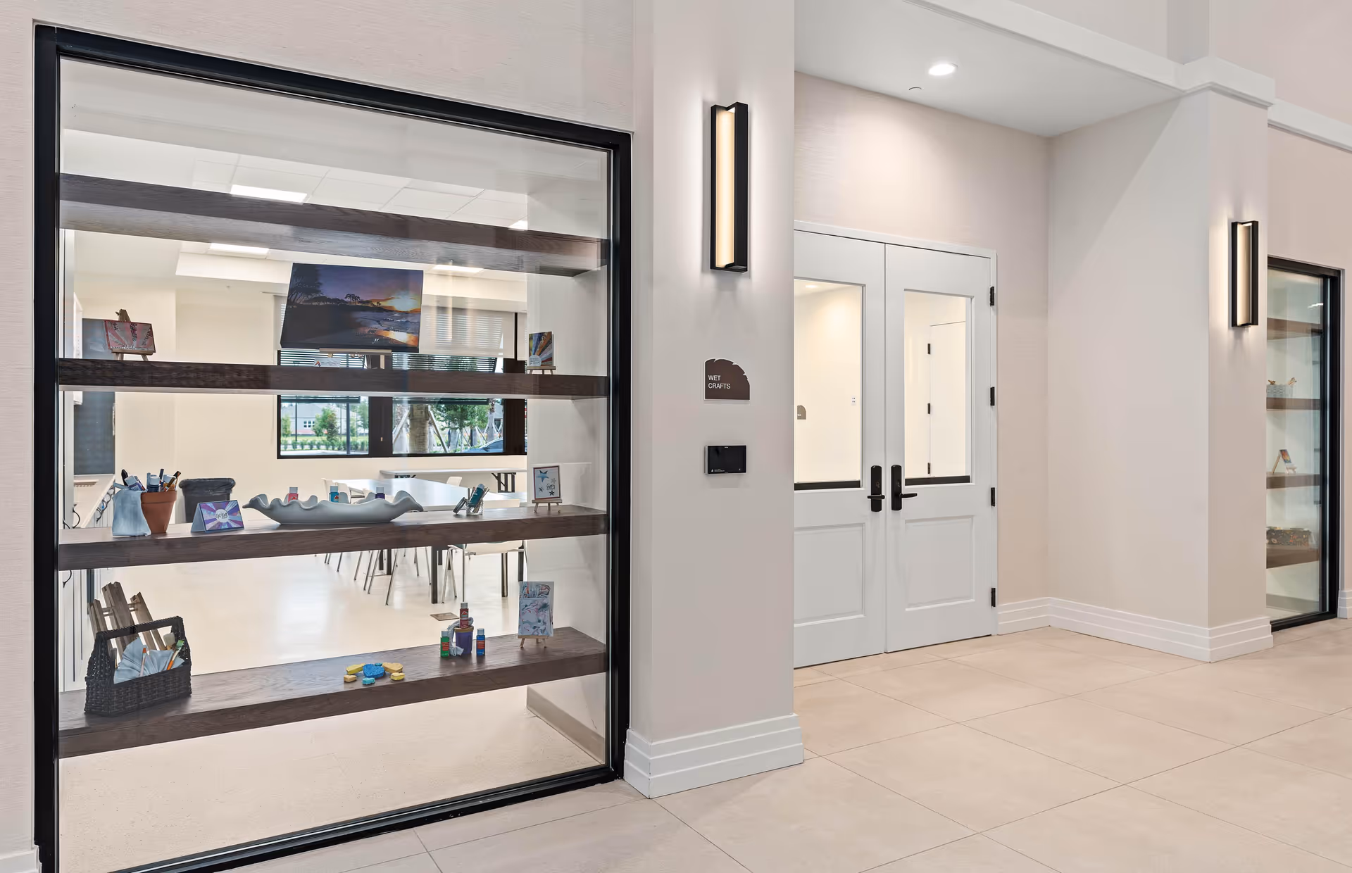 Interior view of a hallway in a senior living facility with large glass windows displaying art and craft items on wooden shelves. Double white doors labeled 'Wet Crafts' lead into a bright room with tables and chairs, and modern wall lights illuminate the space.
