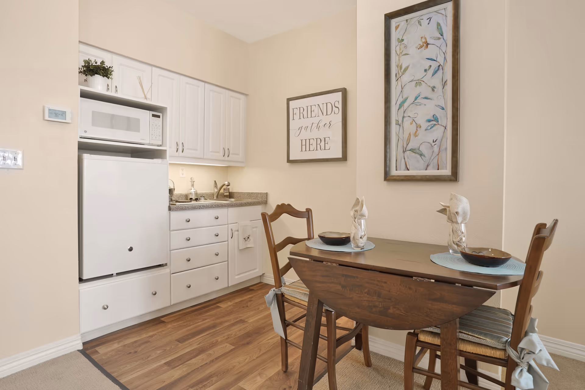 A small dining area with a wooden drop-leaf table set for two, featuring two wooden chairs with cushions. Adjacent to the dining area is a compact kitchen with white cabinets, a microwave, a mini refrigerator, and a granite countertop with a sink. The walls are beige, decorated with a framed floral artwork and a sign that reads 'FRIENDS gather HERE'. The floor is a combination of wood and carpet.