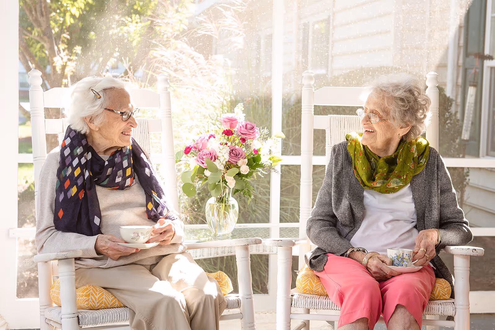 Two elderly women sitting on white rocking chairs on a sunlit porch, smiling and holding teacups. A glass table with a vase of pink and white flowers is between them. Both women wear glasses and colorful scarves.