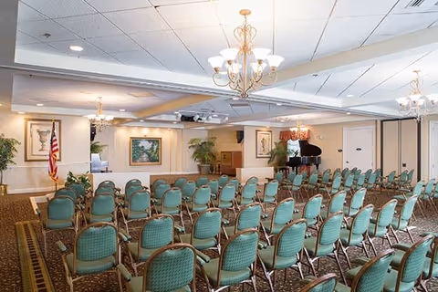 A large meeting or event room with rows of green cushioned chairs arranged facing a small stage area with a grand piano, American flag, framed artwork, and potted plants. The ceiling has recessed lighting and chandeliers.