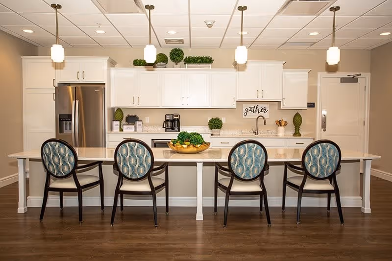 A modern kitchen area with a long white island countertop and four chairs with patterned blue and white backs. The kitchen features white cabinets, a stainless steel refrigerator, a coffee maker, and decorative green plants on the countertop and above the cabinets. Three pendant lights hang from the ceiling above the island. A sign on the wall reads 'gather'.