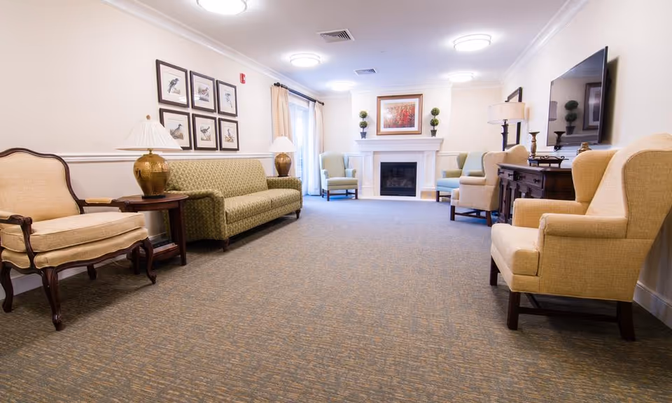 A spacious senior living facility lounge with beige and green upholstered chairs and sofas arranged along the walls. The room features a carpeted floor, a fireplace with decorative plants and a framed painting above it, lamps on side tables, and a large flat-screen TV mounted on the wall.