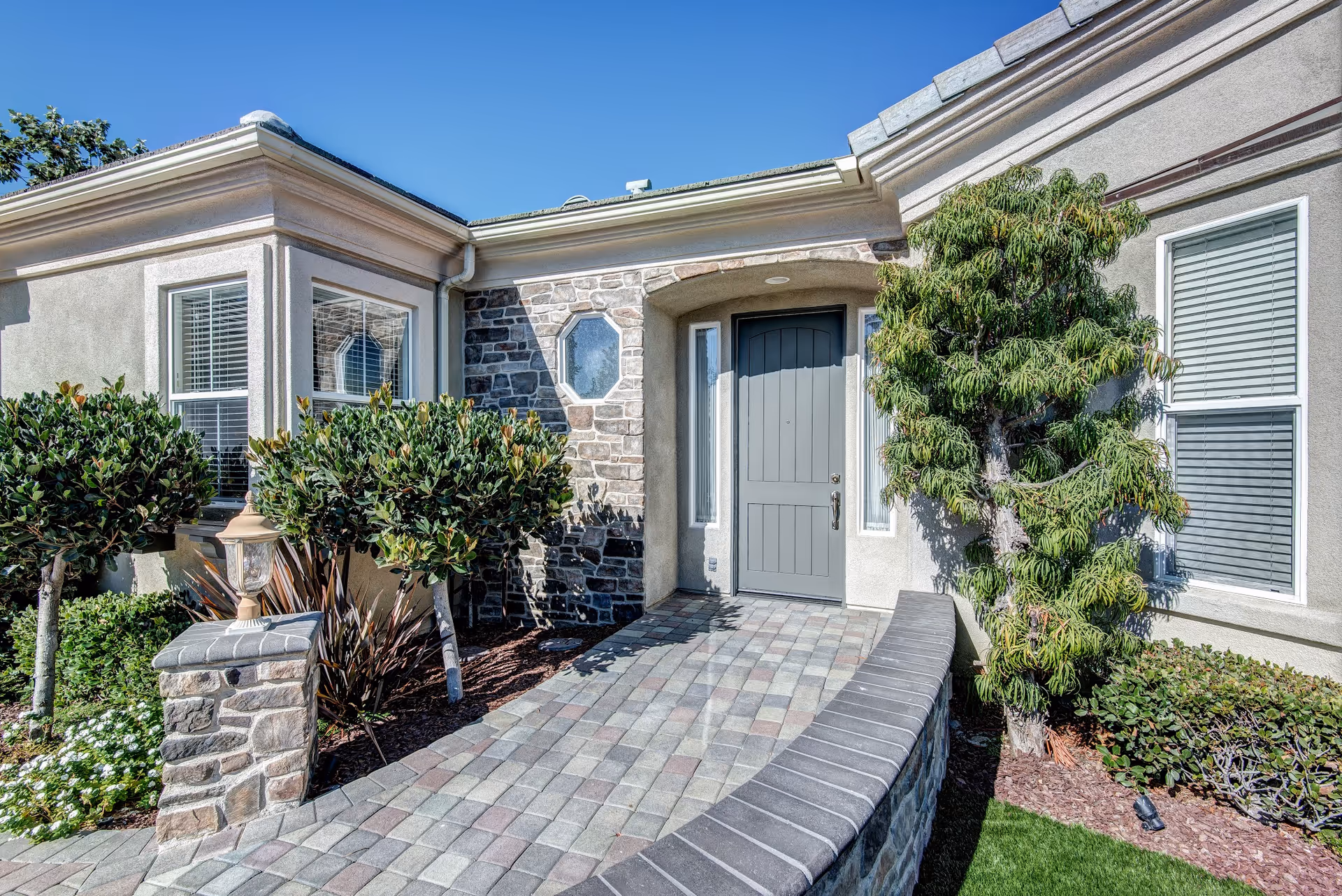 Front entrance of a residential building with a gray door, stone and stucco exterior walls, a paved walkway, and neatly trimmed bushes and trees under a clear blue sky.