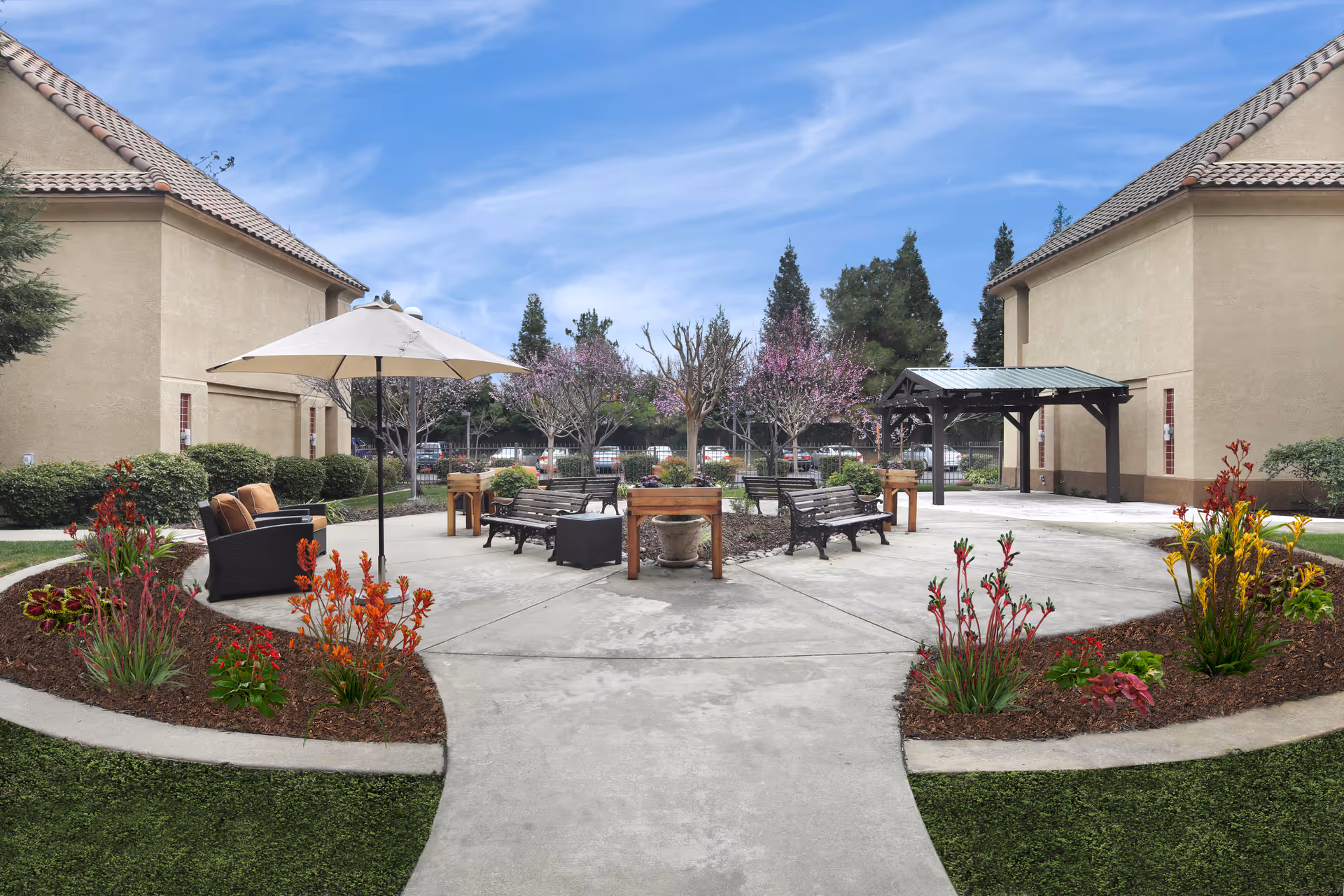 Outdoor courtyard area between two beige buildings with tiled roofs, featuring a circular concrete patio with benches, potted plants, and a large umbrella shading a seating area with cushioned chairs. Flower beds with colorful plants surround the patio, and trees with pink blossoms are visible in the background under a blue sky with wispy clouds.