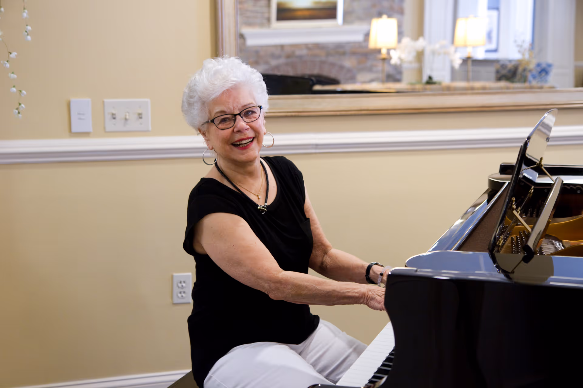 An elderly woman with white hair and glasses is smiling while playing a grand piano in a well-lit room with beige walls and a large mirror reflecting lamps and a fireplace.