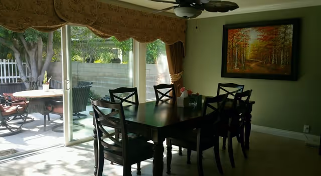 Dining room with a dark wooden table and six matching chairs. A large window with a valance overlooks an outdoor patio area with additional seating and greenery. A ceiling fan is mounted above the table, and a framed painting of a forest scene hangs on the wall.