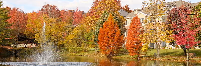A serene outdoor scene at Hill View Health Center featuring a pond with a water fountain in the center, surrounded by trees with vibrant autumn foliage in shades of red, orange, and yellow. A multi-story building is visible in the background behind the trees.