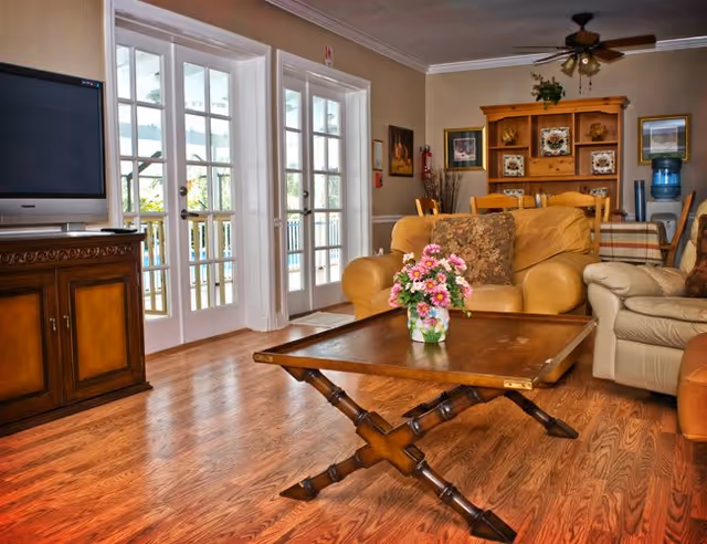 A cozy living room with wooden flooring, a wooden coffee table with a flower vase, two beige sofas with cushions, a wooden TV stand with a flat-screen TV, and a wooden hutch with decorative plates and plants. French doors lead to an outdoor area.
