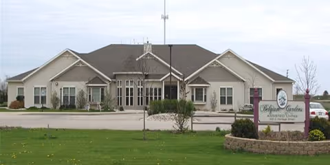 Single-story building with beige siding and multiple gables, surrounded by a paved driveway and green lawn. A sign in front reads 'Belgium Gardens Assisted Living'.