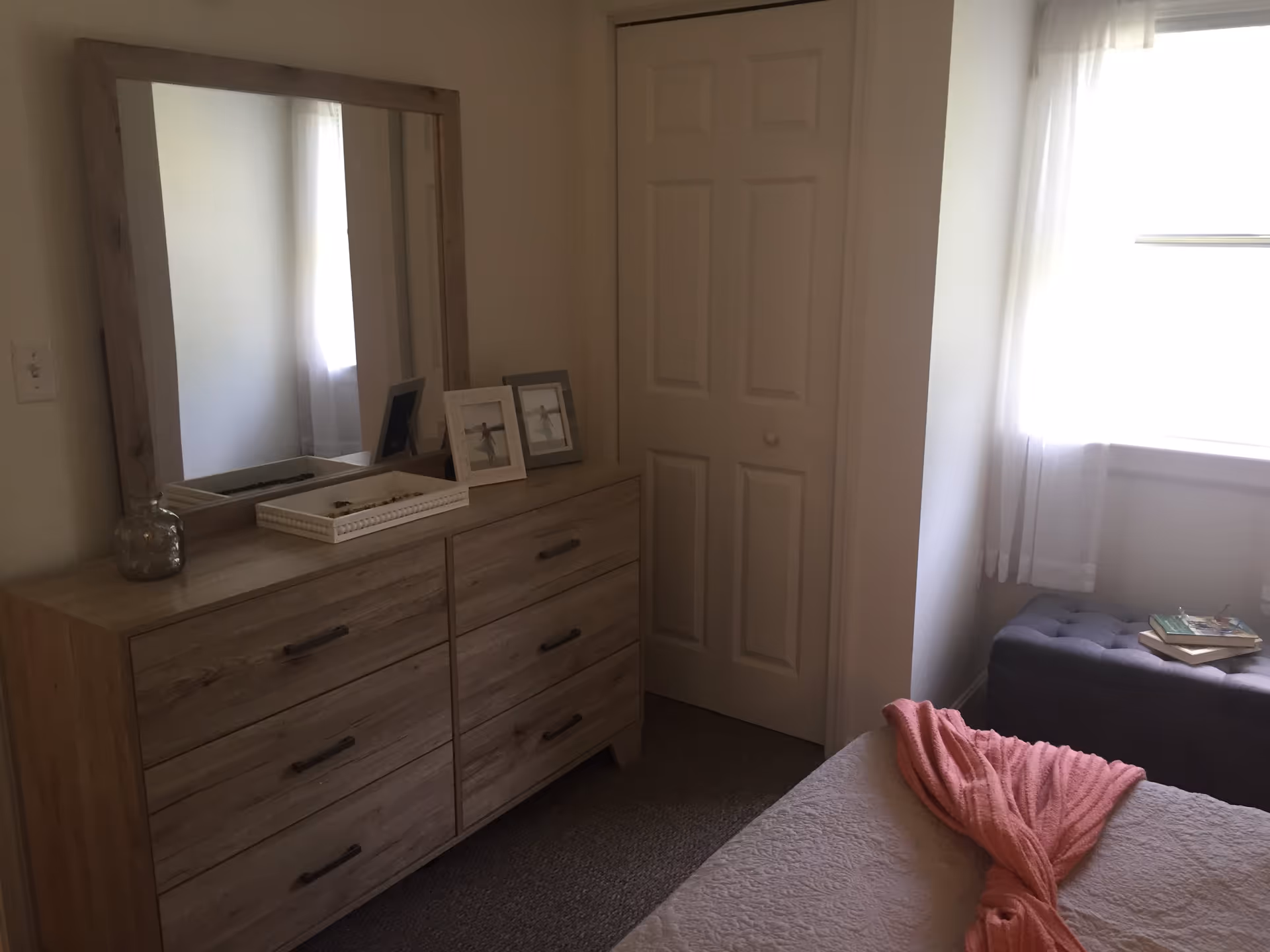 A bedroom corner featuring a wooden dresser with six drawers and a large framed mirror on top. On the dresser are two framed photos, a decorative tray, and a small glass jar. Next to the dresser is a closed white closet door. A window with sheer white curtains lets in natural light, illuminating a gray tufted bench with a few books on it. In the foreground, part of a bed with a white quilt and a pink knitted throw is visible.