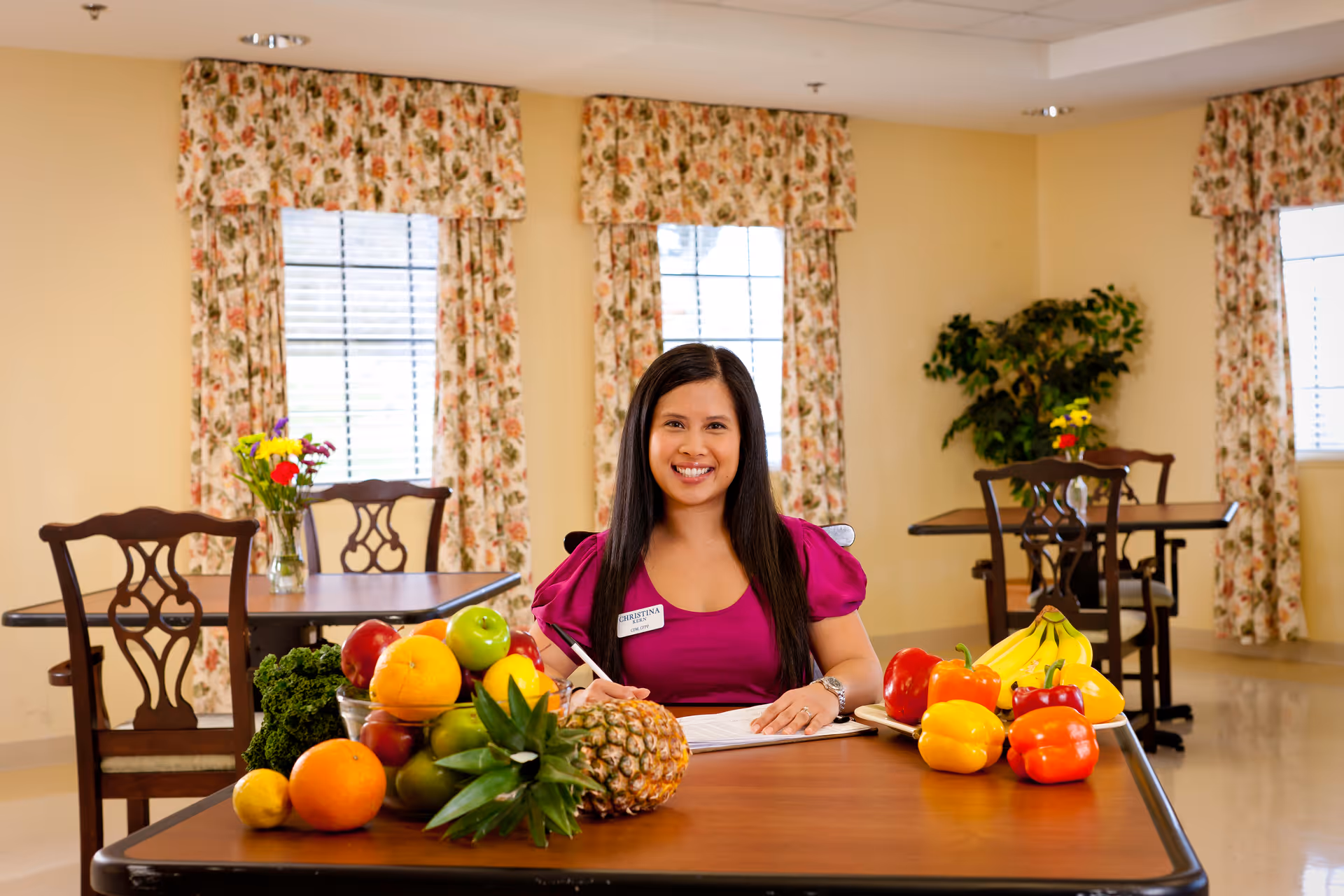 A woman with long dark hair wearing a magenta top sits at a table in a room with floral curtains and several windows. The table is adorned with a variety of fresh fruits and vegetables including a pineapple, bananas, bell peppers, oranges, apples, and leafy greens. The woman is smiling and holding a pen over a notebook.