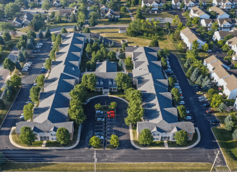 Aerial view of Wellington Village Senior Apartments showing two long residential buildings with pitched roofs, surrounded by trees and parking lots. The complex is situated in a suburban neighborhood with additional houses and greenery visible in the background.