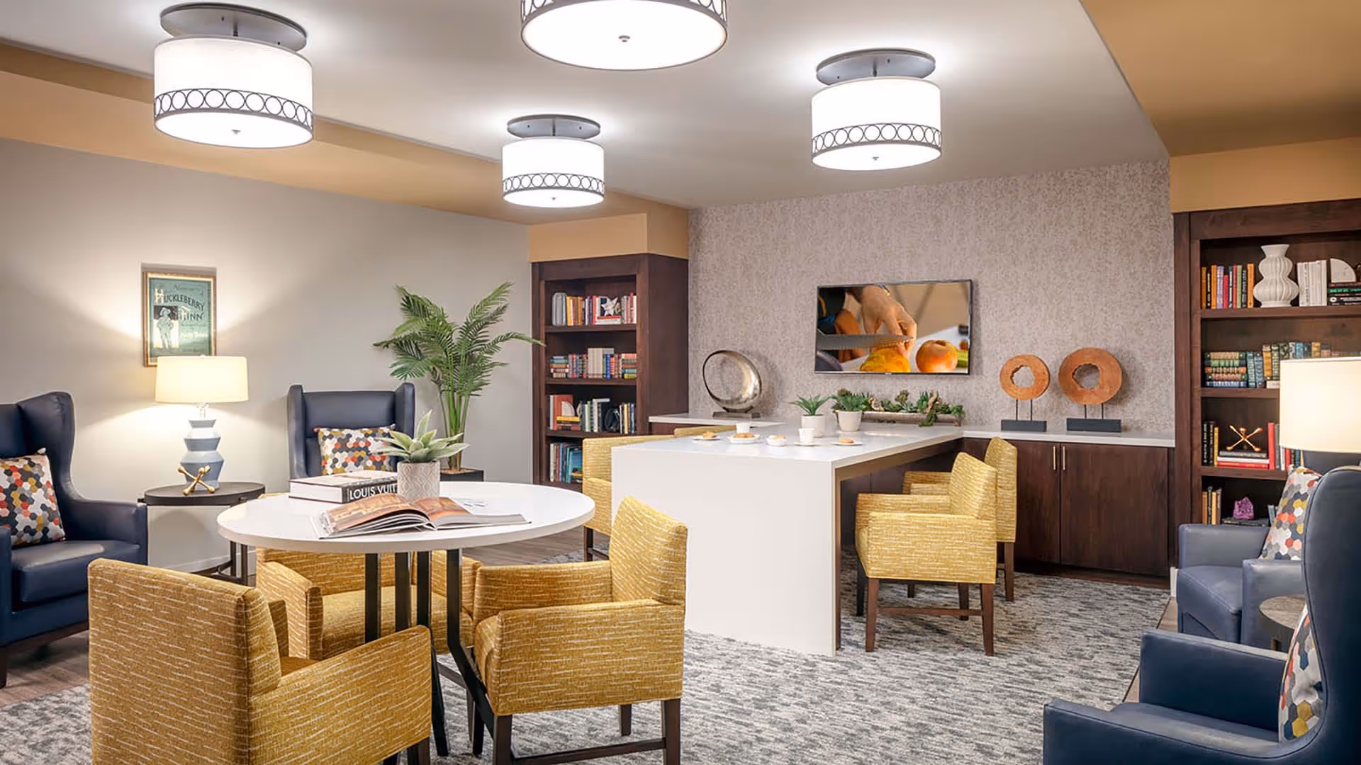 A cozy and well-lit common area with a round white table surrounded by four mustard yellow upholstered chairs. There are two dark blue armchairs with colorful patterned pillows on either side of the room, each next to a small side table with a lamp. The back wall features built-in dark wood bookshelves filled with books and decorative items, and a flat-screen TV mounted above a white counter with more mustard yellow chairs. The room has a soft gray carpet and modern ceiling lights.