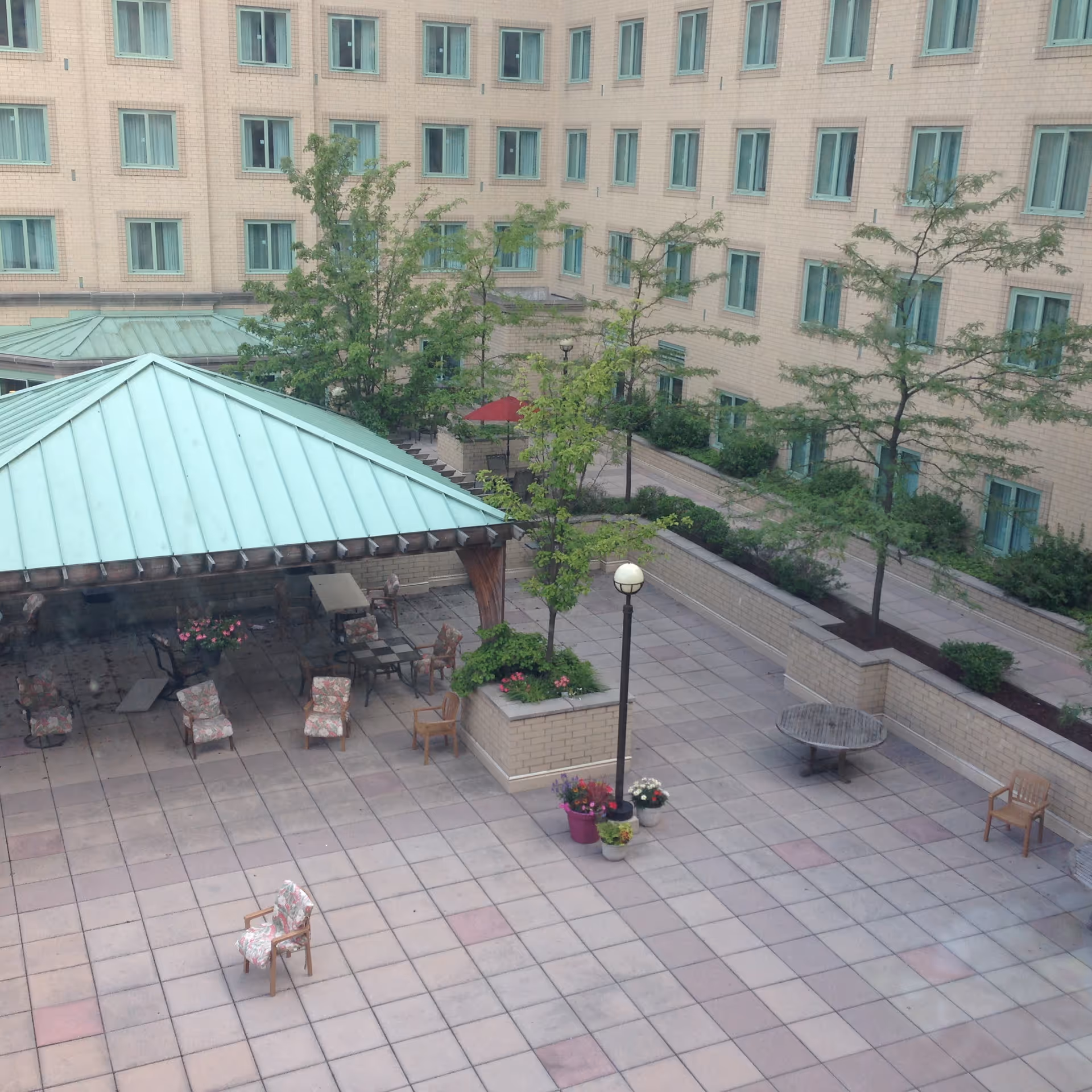 Outdoor courtyard area at Vincentian Schenley Gardens with a covered seating area featuring chairs and tables, surrounded by trees and plants. The courtyard is paved with tiles and enclosed by a multi-story building with many windows.