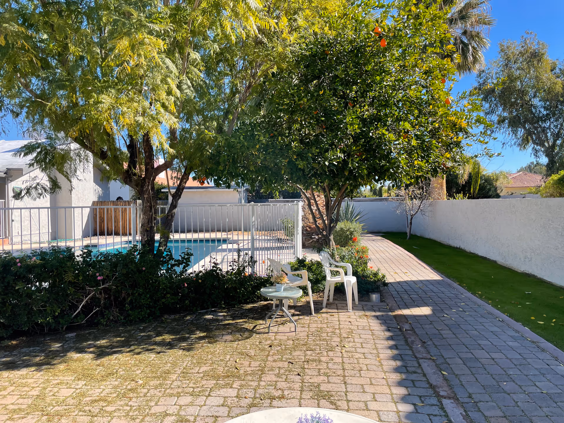Outdoor patio area with a paved walkway, two white plastic chairs, and a small round table under a tree. There is a fenced swimming pool in the background, surrounded by greenery and a white wall enclosing the space. The sky is clear and blue.