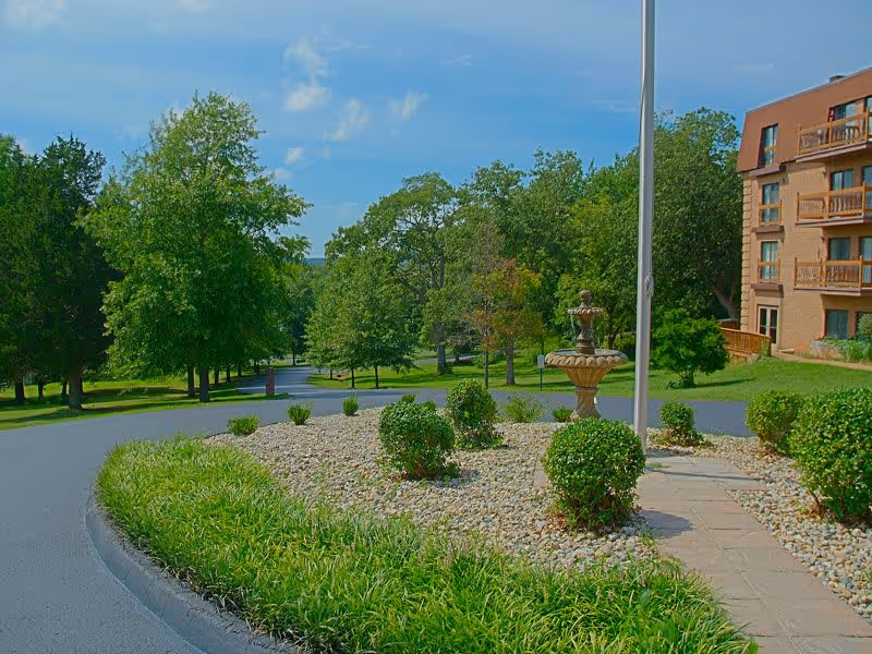 A landscaped outdoor area with a circular rock garden featuring small bushes and a decorative fountain in the center. Surrounding the garden are green trees and a paved road curving around it. A multi-story brick building with balconies is visible on the right side under a blue sky with some clouds.