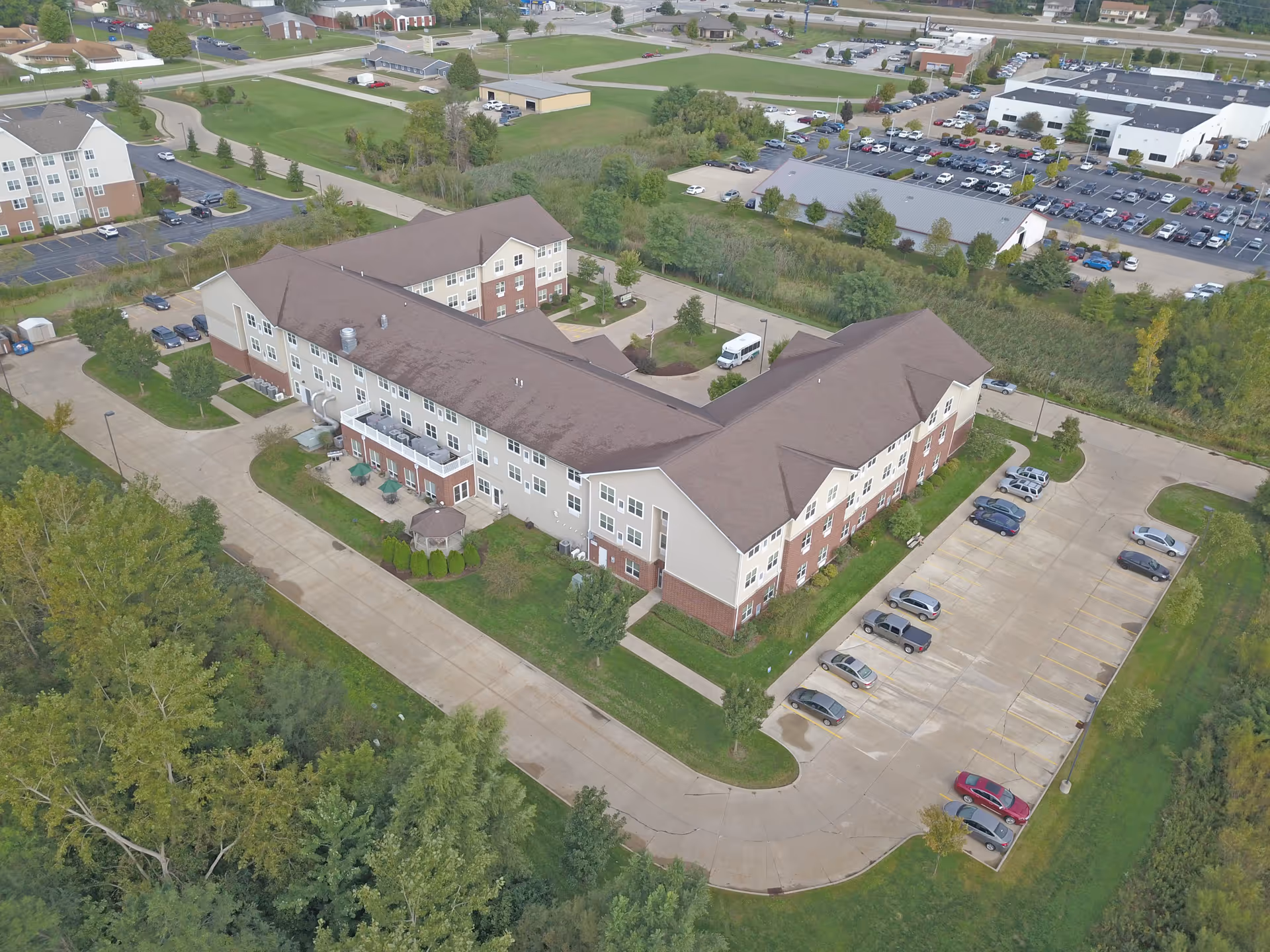 Aerial view of a large senior living facility building with a brown roof and beige and brick exterior walls. The building is surrounded by parking lots with several cars parked, green lawns, trees, and nearby roads. Other buildings and open green spaces are visible in the background.