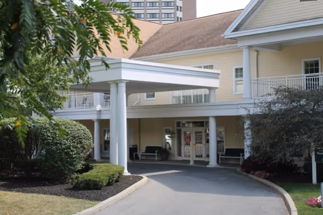 Entrance of a senior living facility with a covered driveway supported by white columns. The building is light yellow with white trim and has a second-floor balcony. There are bushes and trees around the driveway and a tall building visible in the background.