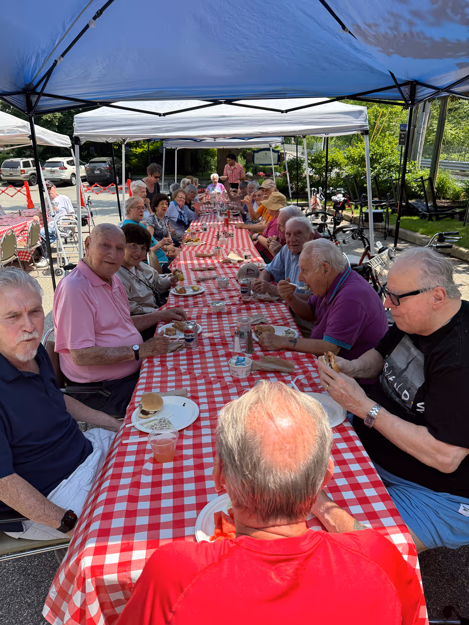 A group of elderly people sitting around a long table covered with a red and white checkered tablecloth under blue and white canopy tents outdoors. They are enjoying a meal together with plates of food and drinks visible on the table. The setting appears to be a social gathering or picnic in a parking lot area with greenery in the background.