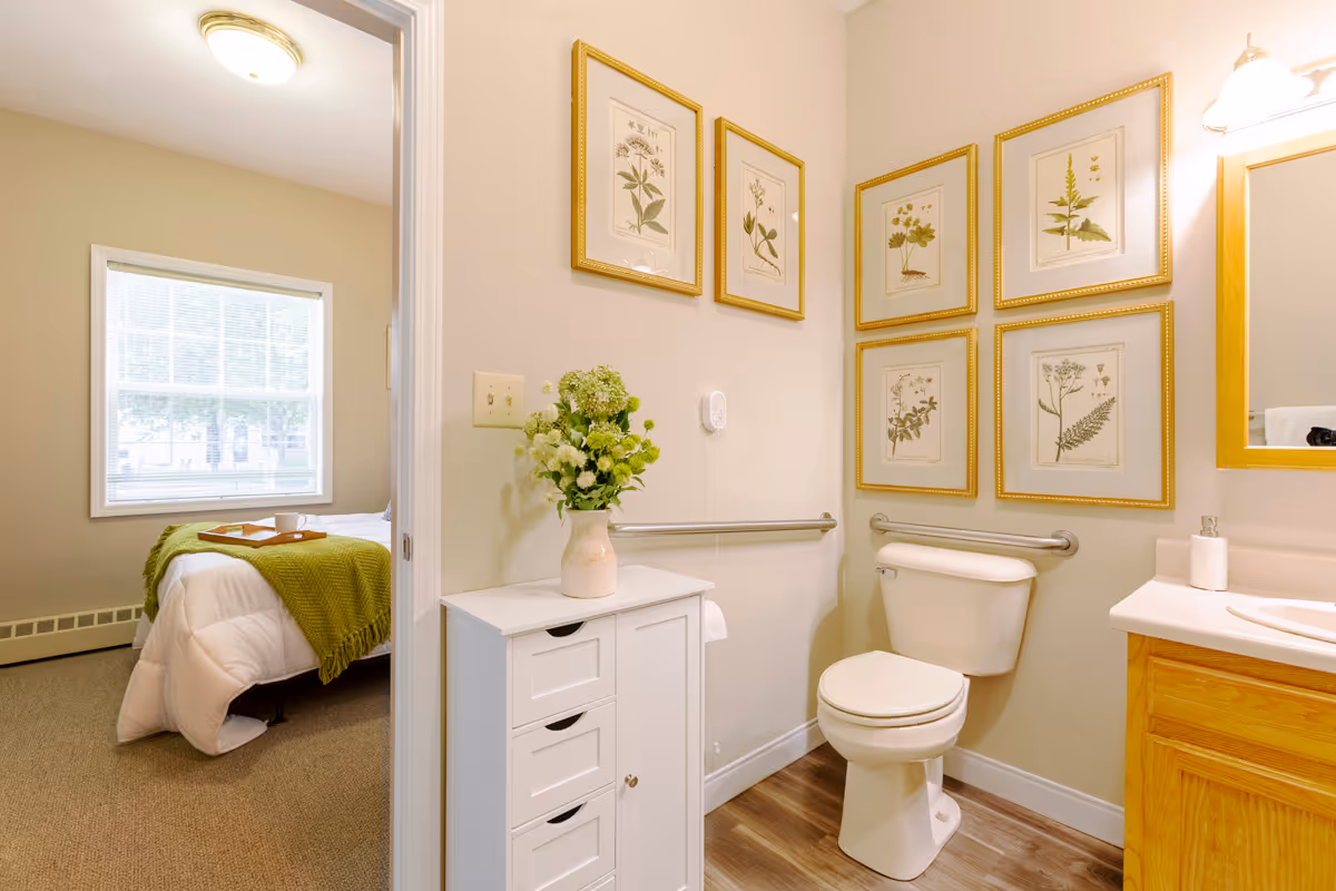 A bright bathroom with a white toilet, wooden vanity with a sink, and a white cabinet topped with a vase of flowers. The walls are decorated with six framed botanical prints. A doorway leads to a bedroom with a bed covered in white bedding and a green throw blanket, with a window letting in natural light.
