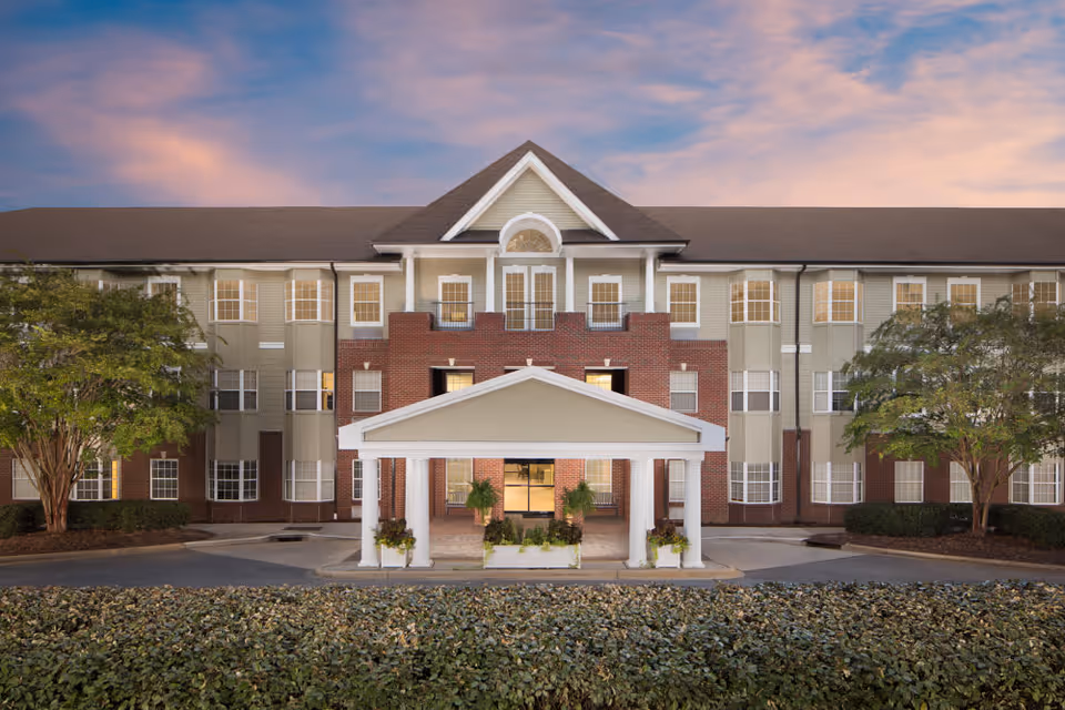 Front exterior view of a three-story senior living facility building with a covered entrance supported by white columns, surrounded by greenery and trees under a colorful sky at dusk.