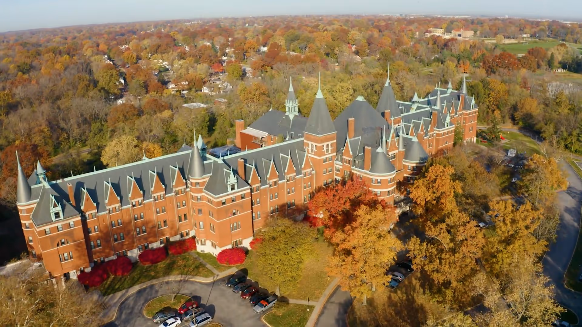 Aerial view of a large, castle-like brick building with multiple pointed towers and steep roofs, surrounded by trees with autumn foliage and a parking area with several cars.