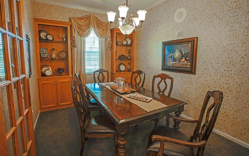 A traditional dining room with a polished wooden table and six matching chairs. The table is decorated with a beige runner, a glass pitcher, and a tray with small items. The room features built-in wooden shelves displaying plates and decorative items, a window with blinds and a swag curtain, a chandelier overhead, and a framed painting on the wallpapered wall.