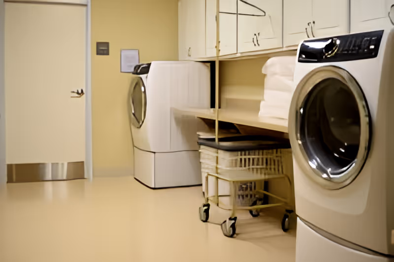 Laundry room with two front-loading washing machines, a laundry cart with wheels, shelves with folded towels, and cabinets above the machines.