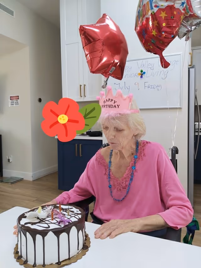 An elderly woman wearing a pink 'Happy Birthday' crown and a pink top sits at a table with a birthday cake decorated with candles and chocolate drizzle. Behind her are red star-shaped and colorful birthday balloons. The background shows a kitchen area with white cabinets and a whiteboard with writing that includes 'Valley Village Senior Living' and the date 'July 9, Friday'.