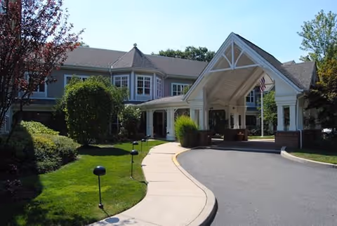 Exterior view of a senior living facility with a covered entrance driveway, landscaped greenery, and a two-story building in the background under a clear sky.