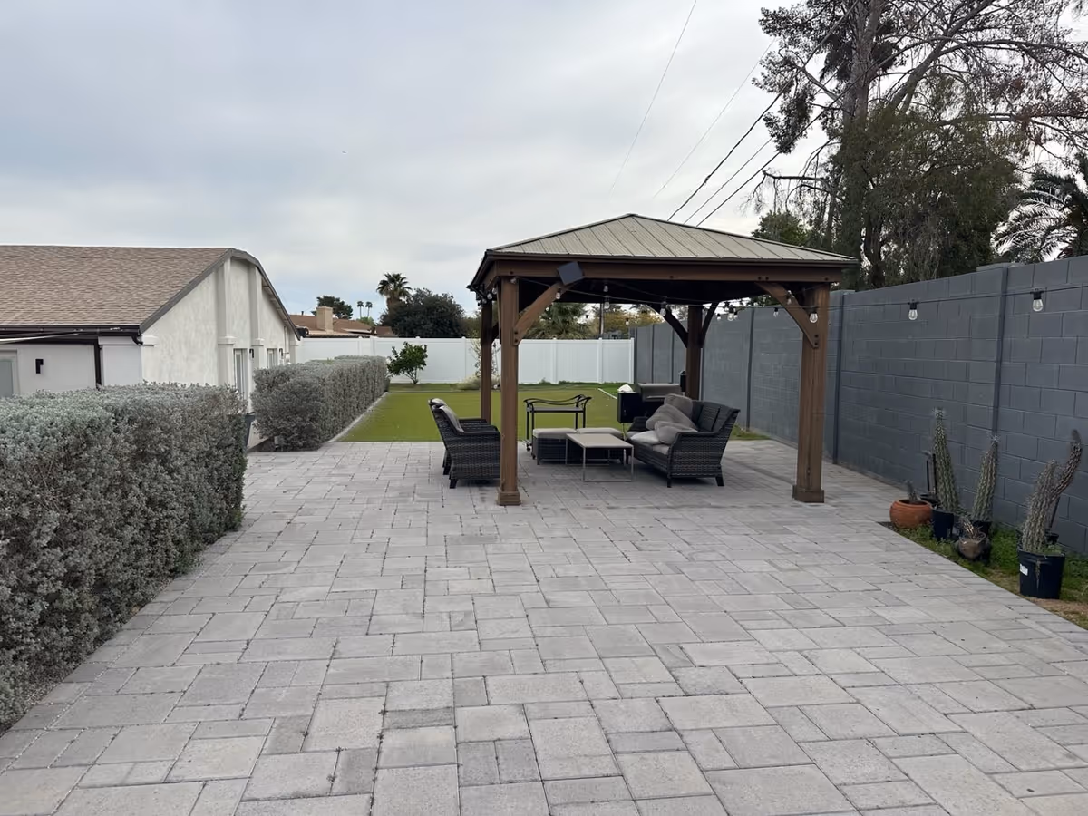 Outdoor patio area with a wooden gazebo covering cushioned wicker furniture including a sofa, chairs, and a coffee table. The patio is paved with gray bricks and bordered by a gray block wall on one side and trimmed bushes on the other. There are string lights hanging on the gazebo and a few potted plants along the wall.