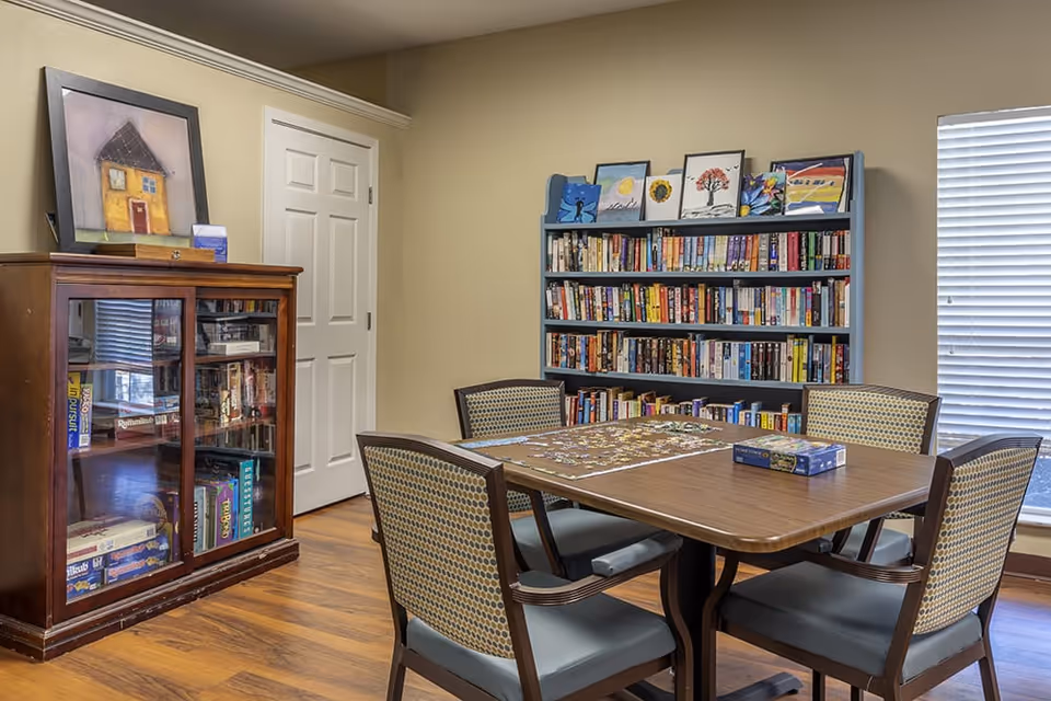 A cozy room with a wooden table and four chairs arranged around it. On the table, there is a partially completed jigsaw puzzle and a puzzle box. Behind the table, there is a blue bookshelf filled with books and several framed artworks displayed on top. To the left, there is a wooden cabinet with glass doors containing board games and books. A closed white door is visible in the background, and a window with blinds is on the right side, allowing natural light into the room. The floor is wooden, and the walls are painted beige.