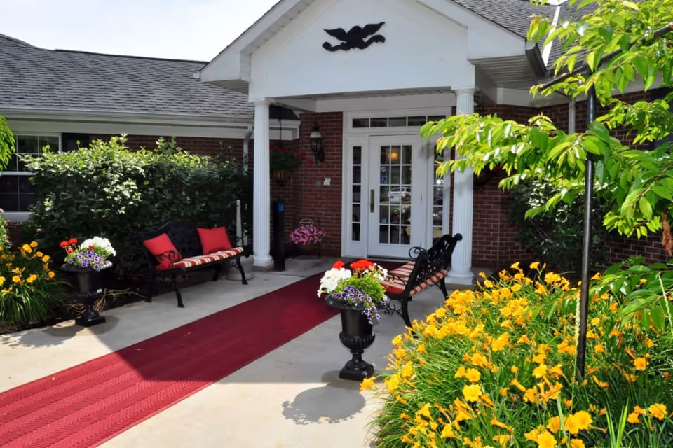 Entrance to a brick building with white columns and a glass door. There are two black benches with red and striped cushions on either side of a red carpet runner leading to the door. The area is surrounded by green bushes and yellow flowers, with potted flowers near the benches.