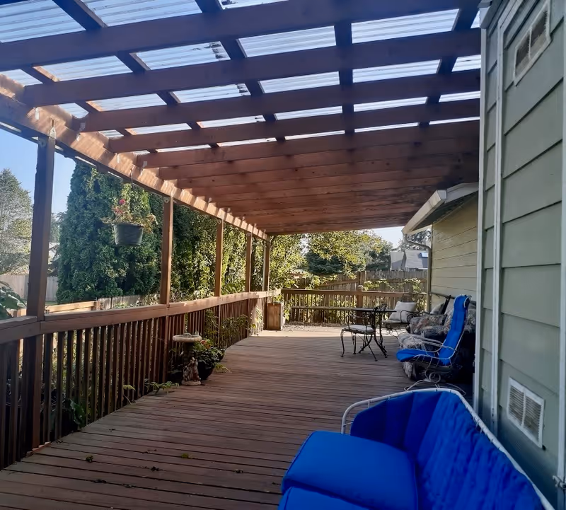 A spacious wooden deck with a pergola overhead, featuring outdoor seating including a blue cushioned chair and a small table with chairs further down the deck. The deck is surrounded by a wooden railing and overlooks a garden area with trees and shrubs.