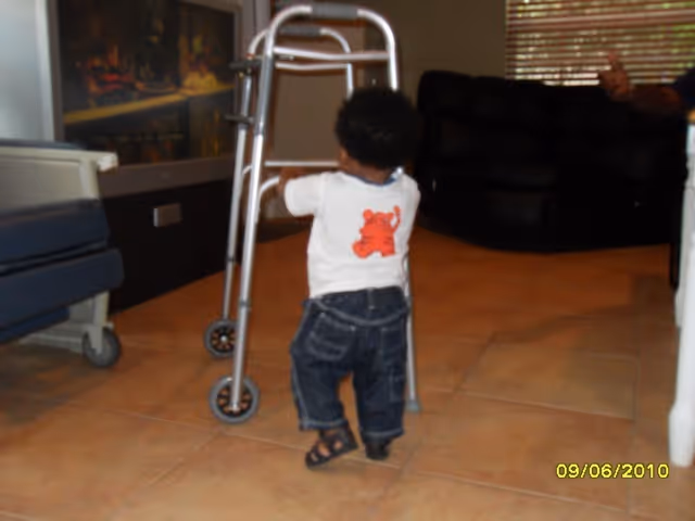 A small child wearing a white shirt with a red bear design and dark jeans is standing and holding onto a metal walker in a room with tiled flooring. In the background, there is a dark couch and a window with blinds.