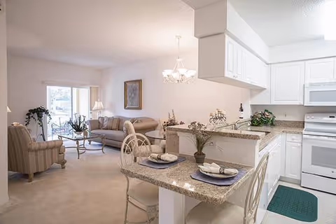 Open-concept living area in a senior living facility featuring a kitchen with white cabinets, granite countertops, and a breakfast bar set with two place settings. Adjacent to the kitchen is a living room with a beige sofa, armchair, glass coffee table, and a sliding glass door leading outside. The space is softly lit with a chandelier above the dining area and a table lamp near the sofa.