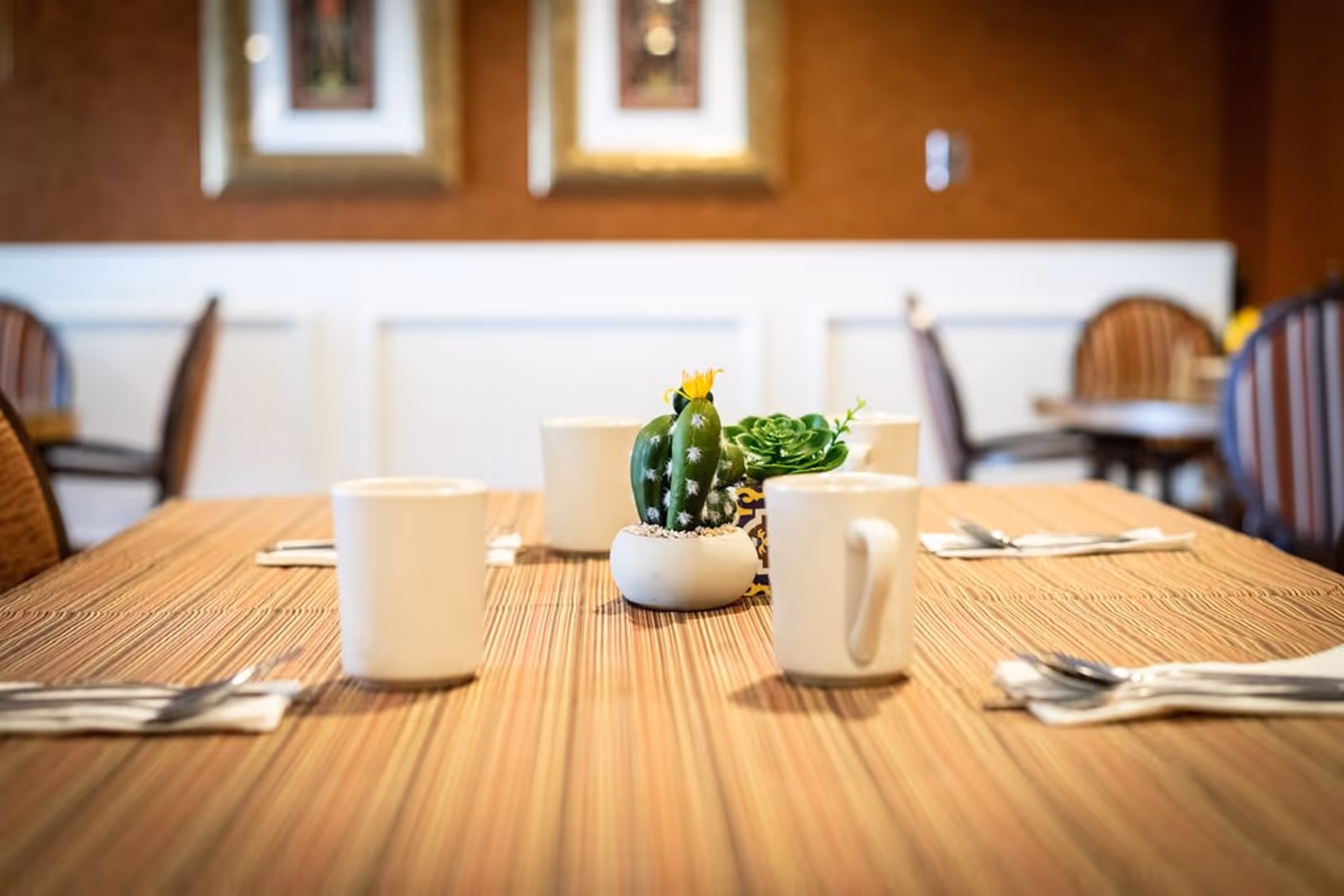 A dining table set with four white mugs, silverware wrapped in napkins, and a small centerpiece featuring a cactus and a succulent plant. The background shows a warm-toned wall with framed artwork and wooden chairs with striped cushions.