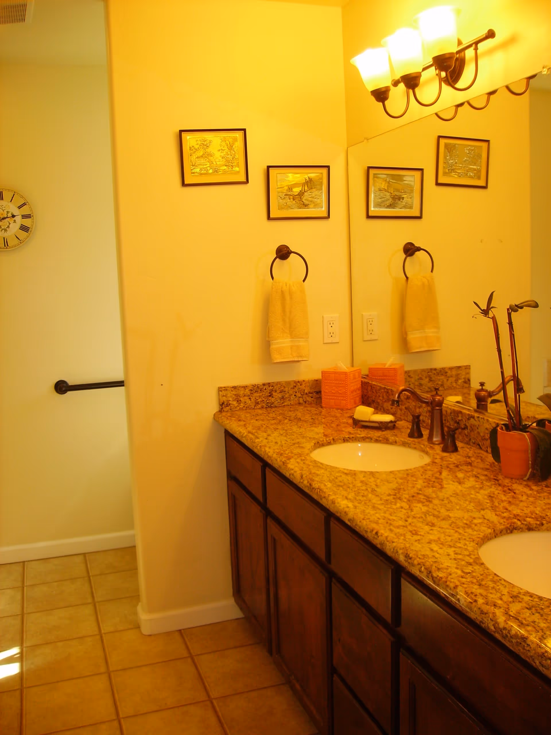 A bathroom with a double sink vanity featuring a granite countertop and dark wooden cabinets. Above the sinks is a large mirror with a three-light fixture. Two framed pictures hang on the wall above a towel ring holding a beige hand towel. The floor is tiled, and a clock is visible on the adjacent wall.
