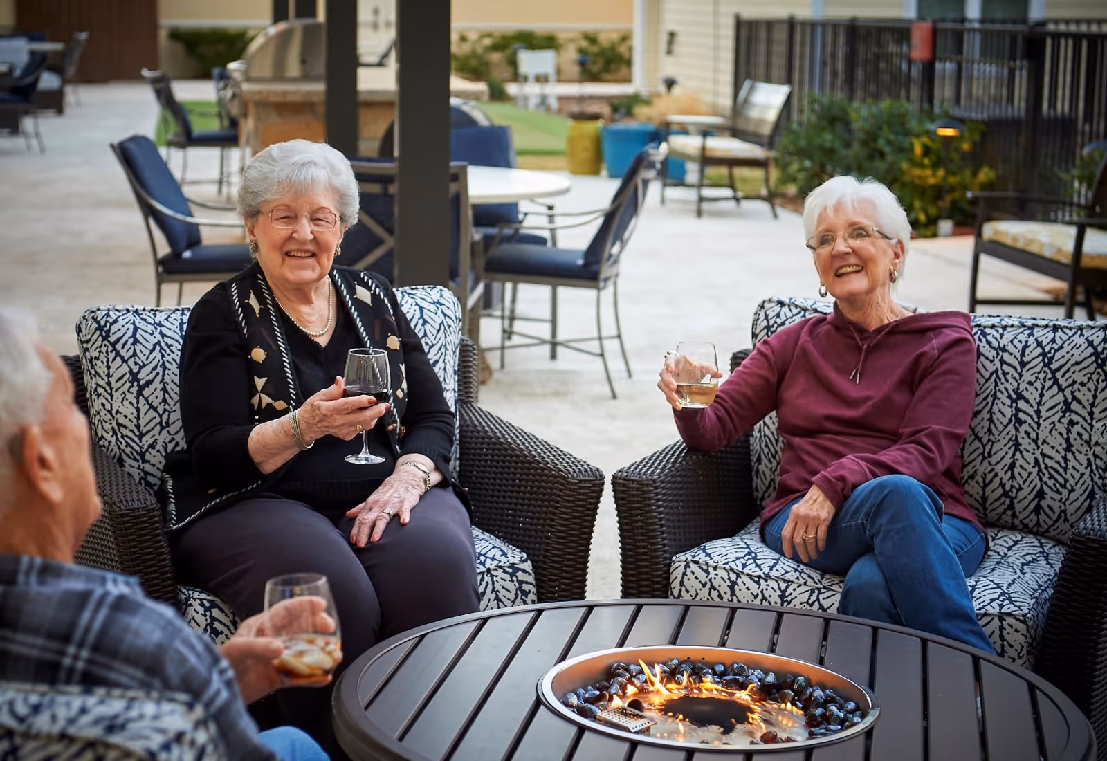 Three elderly people sitting outdoors on cushioned wicker chairs around a round fire pit table, holding drinks and smiling, with outdoor patio furniture and plants in the background.
