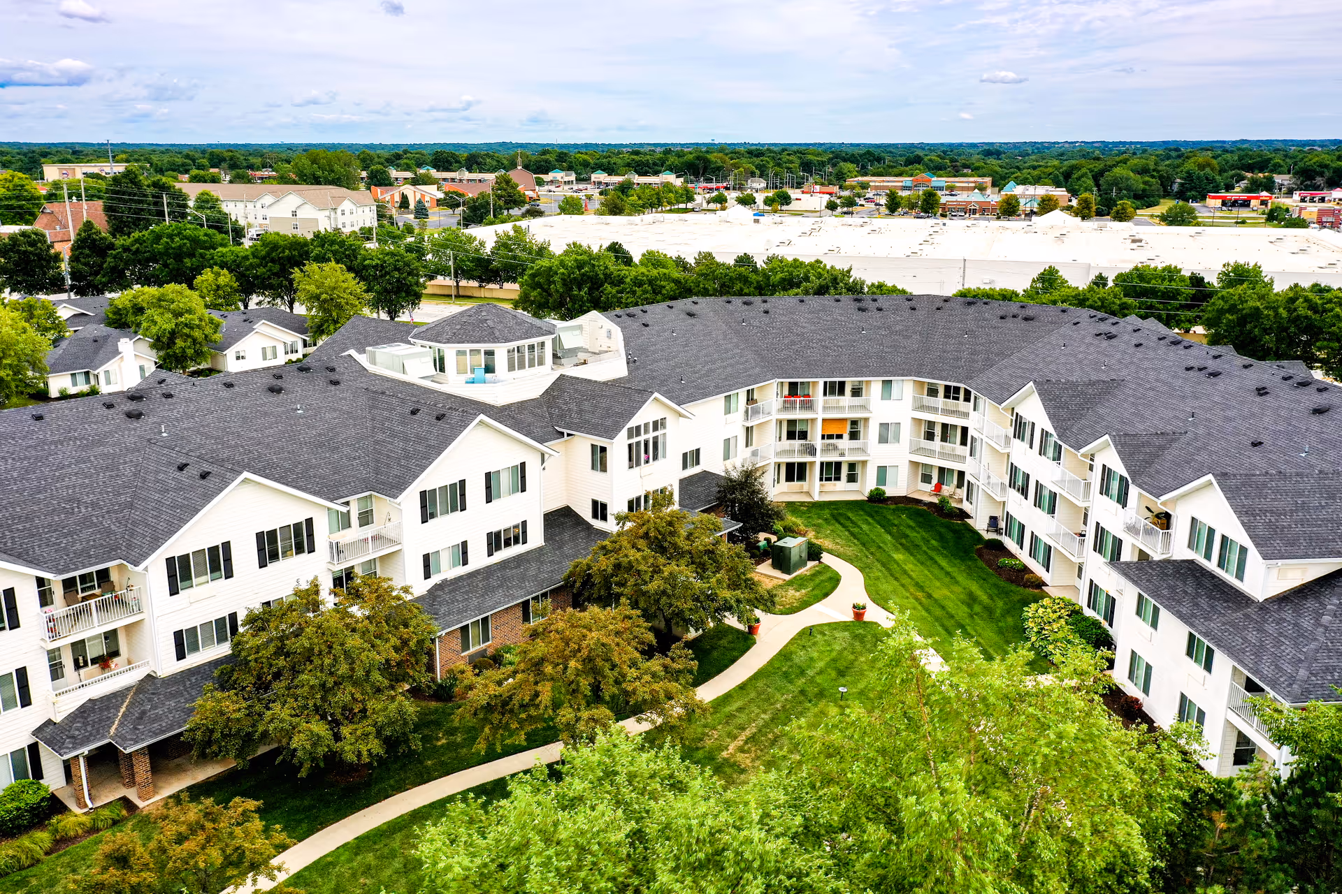 Aerial view of a large senior living facility with white buildings and dark gray roofs surrounding a green courtyard with walking paths and trees. The background shows a suburban area with more buildings and greenery under a partly cloudy sky.