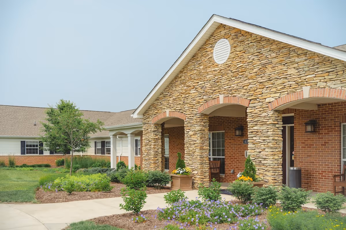 Exterior view of a senior living facility with a stone and brick facade, arched entryways, and a landscaped garden with shrubs and flowers in front.