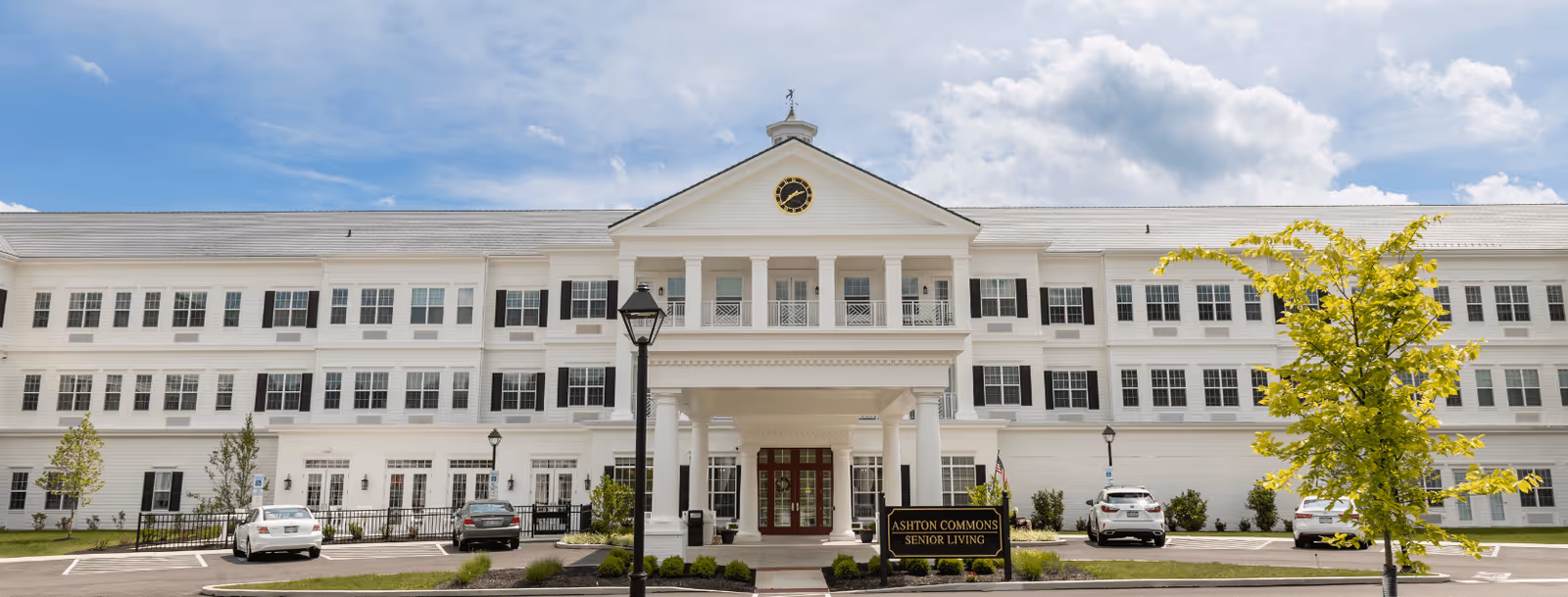 Front exterior view of Ashton Commons Senior Living facility, a large white building with multiple windows, black shutters, a clock on the central pediment, and a covered entrance supported by white columns. Several cars are parked in front, and there are small trees and landscaping around the entrance.