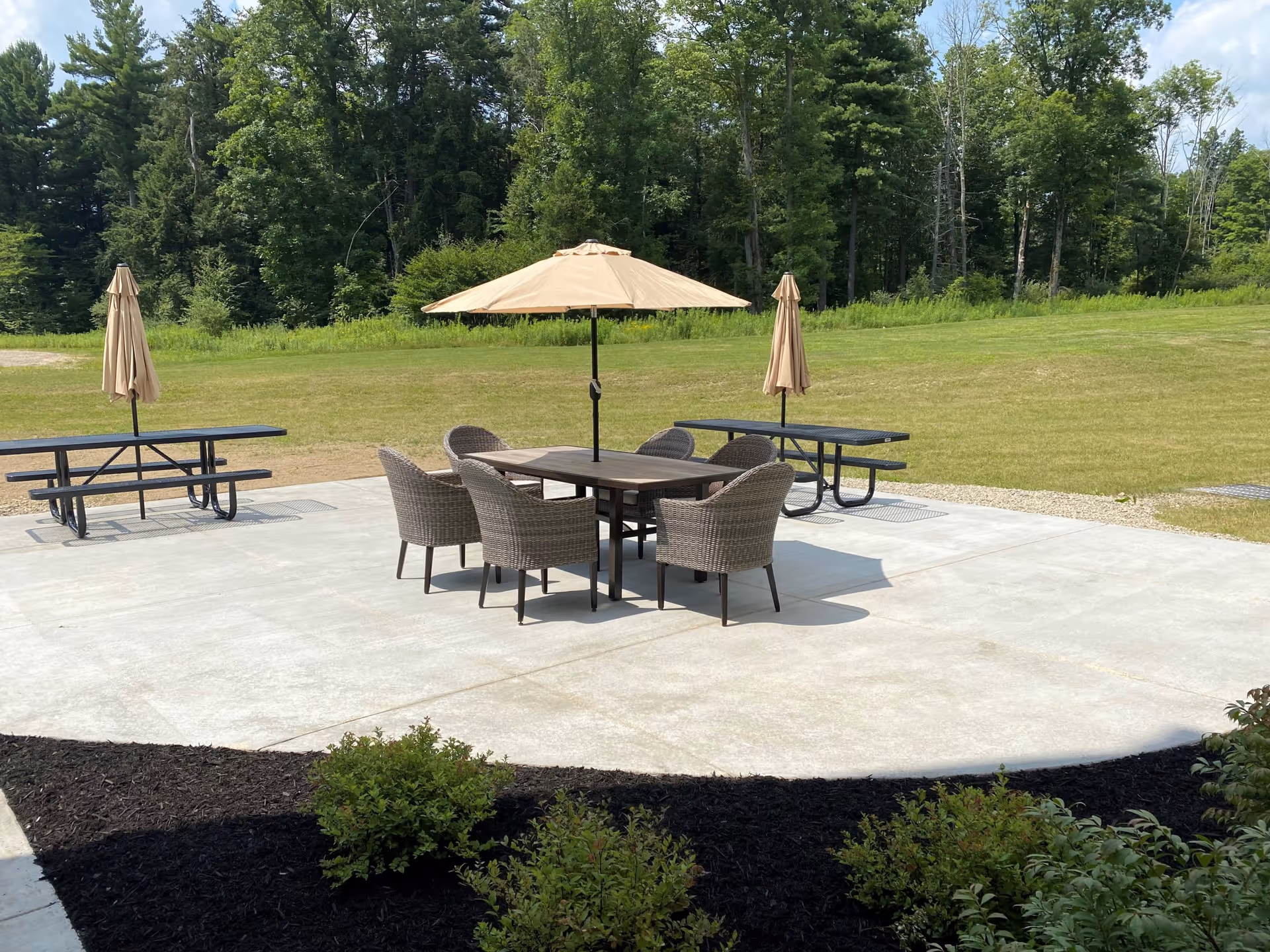Outdoor patio area with a round table surrounded by six wicker chairs and a large beige umbrella in the center. Two picnic tables with closed beige umbrellas are positioned on the concrete patio. The patio is bordered by a grassy lawn and dense trees in the background.