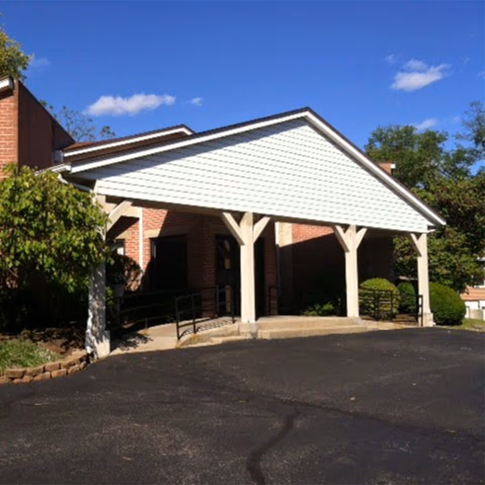 Entrance of a brick building with a white covered porch supported by wooden beams, surrounded by greenery and a clear blue sky.