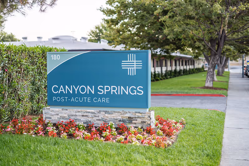 Outdoor view of a sign for Canyon Springs Post-Acute Care, surrounded by green grass and colorful flowers, with a sidewalk and trees in the background.